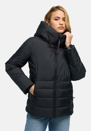Femme blonde portant une doudoune noire avec col montant et fermeture éclair, debout avec une main dans la poche sur un fond uni.