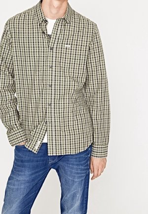 Uomo che indossa una camicia a quadri verde e beige sopra una T-shirt bianca con jeans blu, in piedi contro uno sfondo bianco uniforme.