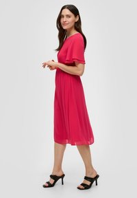 Knielanges rosa Chiffonkleid mit tailliertem Bund und fließenden Ärmeln, kombiniert mit schwarzen Open-Toe-Pumps. Schlichtes, fließendes Design.