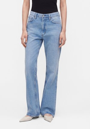 Femme portant un jean bleu clair taille haute à jambes droites, un top noir sans manches et des chaussures gris clair à bout pointu, debout sur fond blanc.