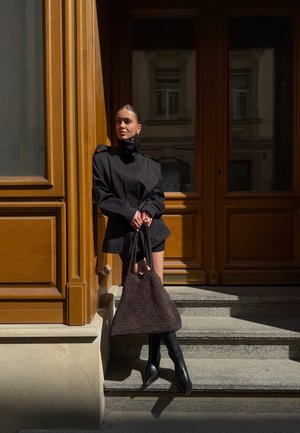 Frau in schwarzer Jacke, Shorts und Stiefeln steht auf Stufen neben einem holzverkleideten Gebäude und hält eine große strukturierte dunkle Handtasche im Sonnenlicht.