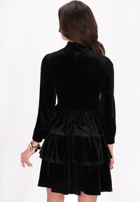 Fekete bársonydressz hosszú ujjakkal, magas nyakkal és tiered fodros kialakítással a derékon, sima textúrával és finom fényességgel.