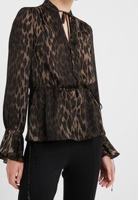 Blusa con stampa leopardata in marrone scuro e nero, dotata di collo a cravatta, maniche lunghe con polsini svasati e vita regolabile con cordoncino.