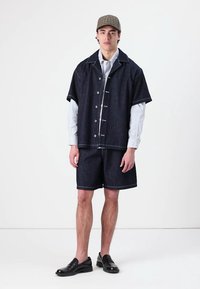 Jeansskjorte med korte ermer, knaplukning og kontrasterende syninger, kombinert med mørke denimshorts og svarte sko.