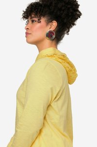 Žlutá mikina s kapucí zdobenou krajkou, vyrobená z jemného, lehkého materiálu s volným střihem. Velké kulaté náušnice doplňují design.