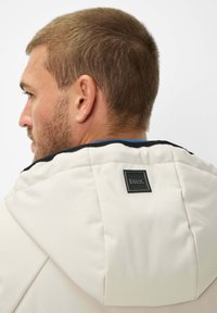 Giacca beige con colletto alto e dettaglio nero sulla spalla. Presenta un'etichetta nera rettangolare sul retro. Texture liscia.