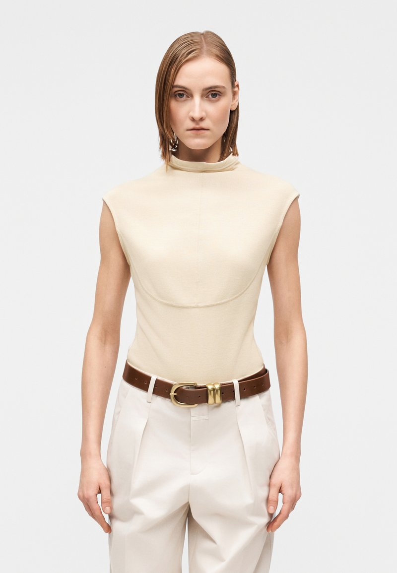 Beige ribbad topp med hög hals och ärmlösa capärmar, kombinerad med högmidjade cremefärgade byxor och ett brunt bälte med guldfärgad spänne.