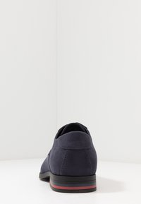 Chaussure en daim marine au design épuré, dotée d'un bout arrondi et d'une semelle rayée rouge et bleue contrastante. Texture lisse et minimalisme dans les détails.