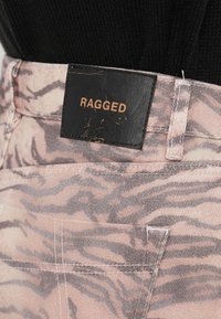 Pink patterned denim pants feature a black leather label with "RAGGED" embossed in gold. Design includes visible stitching and a textured surface.