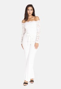 Hvid blonde off-shoulder top med lange ærmer, knyttet i taljen, og tætsiddende hvide bukser. Model iført sorte sandaler med åbne tæer.