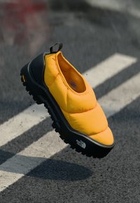 Chaussure jaune en toile du type slip-on avec une semelle noire texturée. Dispose d'un dessus rembourré et d'une languette à l'arrière. Marquage sur le côté.