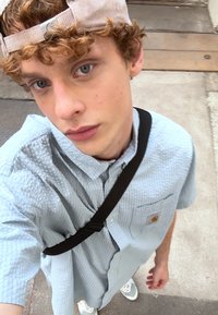 Chemise à boutons texturée bleu clair à manches courtes, sac à bandoulière noir et casquette beige. Une tenue décontractée avec des baskets blanches.