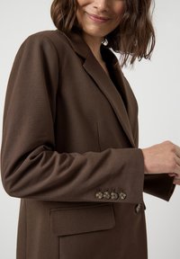 Blazer marrone su misura con rever a scaglie, tessuto testurizzato, chiusura con un bottone e cinque bottoni decorativi sulla manica.