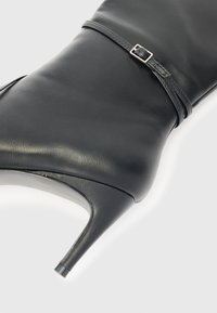 Botín de cuero negro con tacón alto delgado y una correa fina con hebilla de plata alrededor del tobillo.