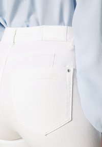 Jeans in denim bianco con una discreta tasca posteriore, caratterizzati da cuciture sottili, una finitura texture e un'etichetta in pelle bianca semplice.