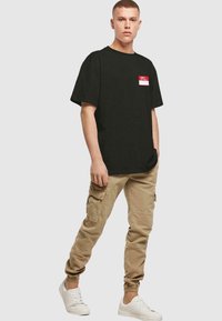 Camiseta negra de manga corta con un logo rojo y blanco en el bolsillo del pecho izquierdo, combinada con pantalones cargo color crema y zapatillas blancas.