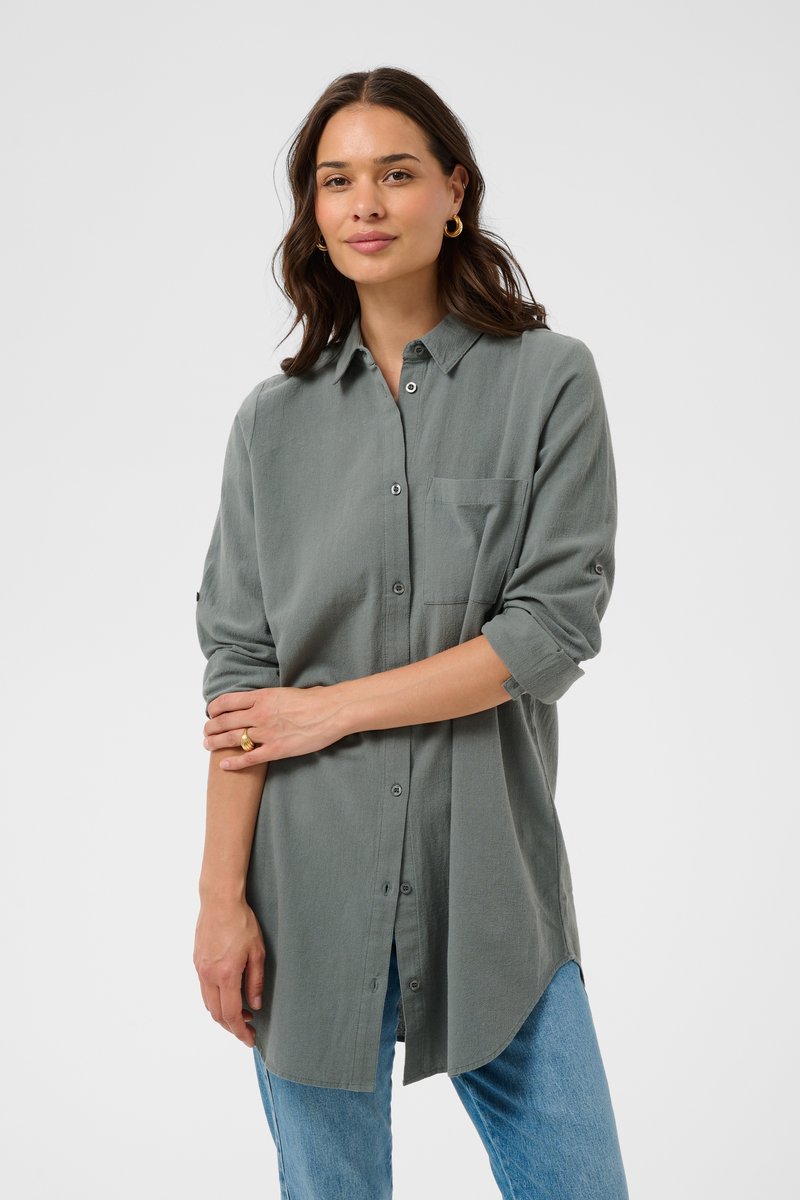 Donna con capelli medio scuri che indossa orecchini a cerchio d'oro, una lunga camicia grigia con bottoni e maniche arrotolate, e jeans blu, in piedi con un braccio incrociato sul corpo.
