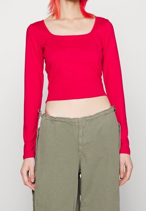 Rotes langärmliges Cropped-Top mit quadratischem Ausschnitt, kombiniert mit lockeren olivgrünen Jogginghosen mit Kordelzug. Glatte Stoffoberfläche, figurbetontes Design.