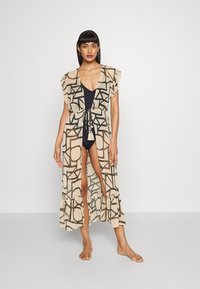 Lange beige cover-up met zwarte geometrische patronen, korte mouwen en een gerimpelde zoom. Draag het over een zwart zwemkostuum, met een trekkoord in de taille.