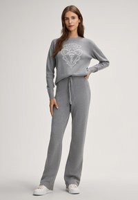 Grijze sweatshirts en bijpassende broekset gemaakt van zachte stof. Heeft een logo op de borst, een relaxte pasvorm en een trekkoord in de taille. Witte sneakers.