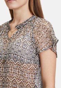 Doorzichtig, bruine blouse met geometrische patronen, geraffineerde kraag, korte mouwen en een keyhole-halslijn verfraaid met een gouden ketting.