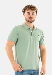 Polo de manga corta verde claro hecho de algodón. Presenta una tapeta de tres botones y un pequeño logotipo en el lado izquierdo del pecho. Textura lisa.