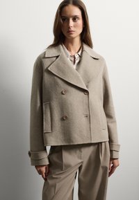Chaqueta de lana beige de corte cruzado con grandes solapas, dos botones y bolsillos laterales. Presenta un ajuste relajado y mangas con puños.