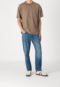 Ruskea puuvillapaita, jossa on lyhyet hihat, pyöreä pääntie ja näkyvät saumat, yhdistettyinä sinisiin suoralahkeisiin farkkuihin ja beigeihin tennareihin, joissa on tummia yksityiskohtia.