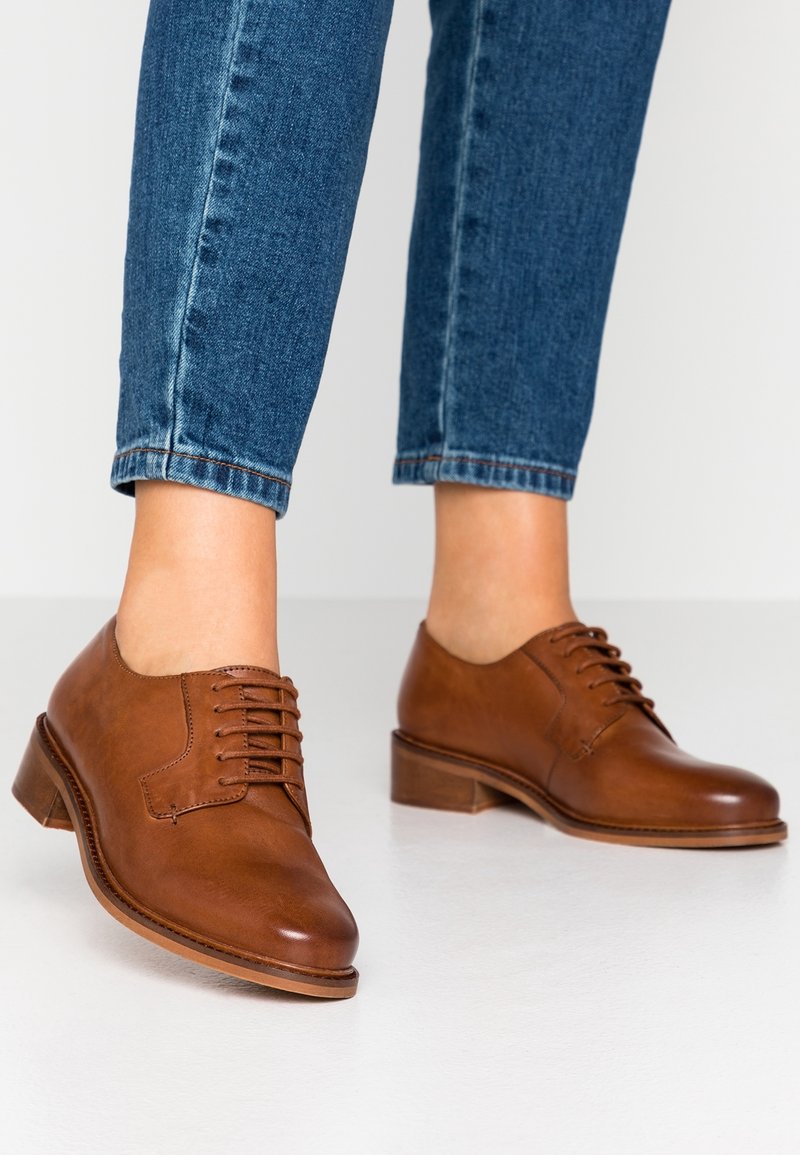 Personne portant un jean bleu court et des chaussures oxford marron en cuir à lacets, debout sur un sol blanc.
