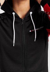 Felpa nera con zip e cordini bianchi, decorata con dettagli rossi, con il logo "Tommy Sport" sul petto, indossata da una persona che sta sistemando il cappuccio.