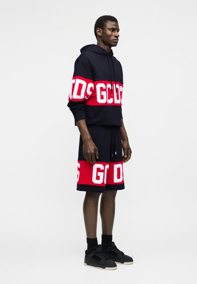 Άνδρας που στέκεται φορώντας μαύρη και κόκκινη μπλούζα με κουκούλα και ταιριαστά σορτς με λευκά έντονα γράμματα και μαύρα αθλητικά παπούτσια μπροστά σε απλό φόντο.