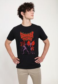 Czarny bawełniany t-shirt z kolorowym nadrukiem "Stranger Things" przedstawiającym potwory w odcieniach fioletu i czerwieni. Krótkie rękawy i okrągły dekolt.