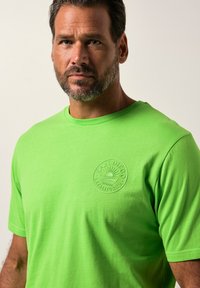 T-shirt di cotone verde con collo a giro e logo testurizzato in design circolare nella zona sinistra del petto. Maniche corte.