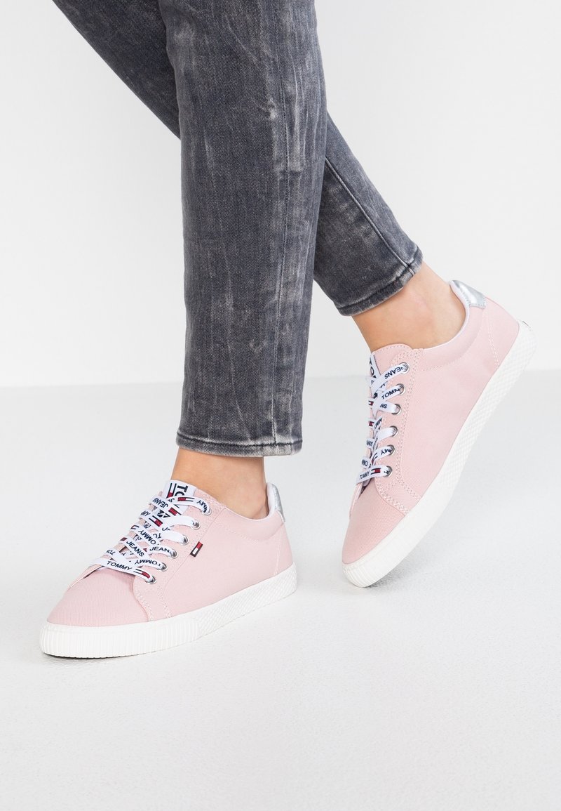 Rosa canvas sneakers med vita gummisulor, metalliskt silverfärgade detaljer vid hälen och mönstrade snören med varumärkeslogotyp. Bärs med grå jeans.