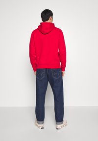 Sudadera roja con capucha ajustable, combinada con jeans oscuros con costuras amarillas en los bolsillos traseros y puños enrollados.