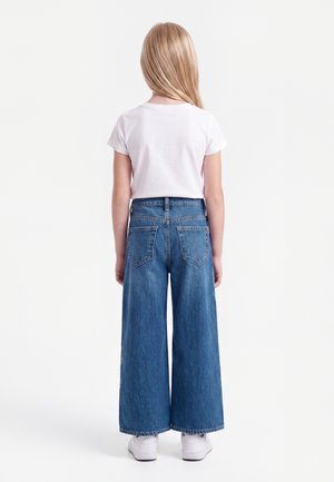 Barn stående med ryggen mod kamera iført en hvid kortærmet t-shirt, blå vide jeans og hvide sneakers på en ensfarvet hvid baggrund.