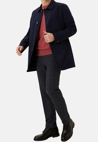 Hombre con un abrigo azul marino, suéter color óxido, pantalones de cuadros oscuros y zapatos de vestir de cuero negro frente a un fondo claro y liso.