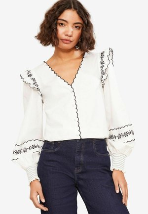 Blusa bianca con ricami neri e bordi scallop, con maniche lunghe voluminose e scollatura a V. Abbinata a jeans in denim scuro.