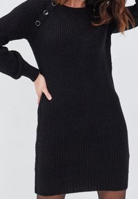 Robe pull noire en tricot avec texture côtelée, présentant des accents de matériel au niveau des épaules et des manches longues. Coupe ajustée avec un col rond.
