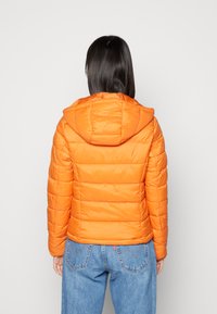 Casaco puffer laranja com design acolchoado horizontal, capuz e textura suave. Usado sobre jeans de denim azuis, mostrando a vista traseira.
