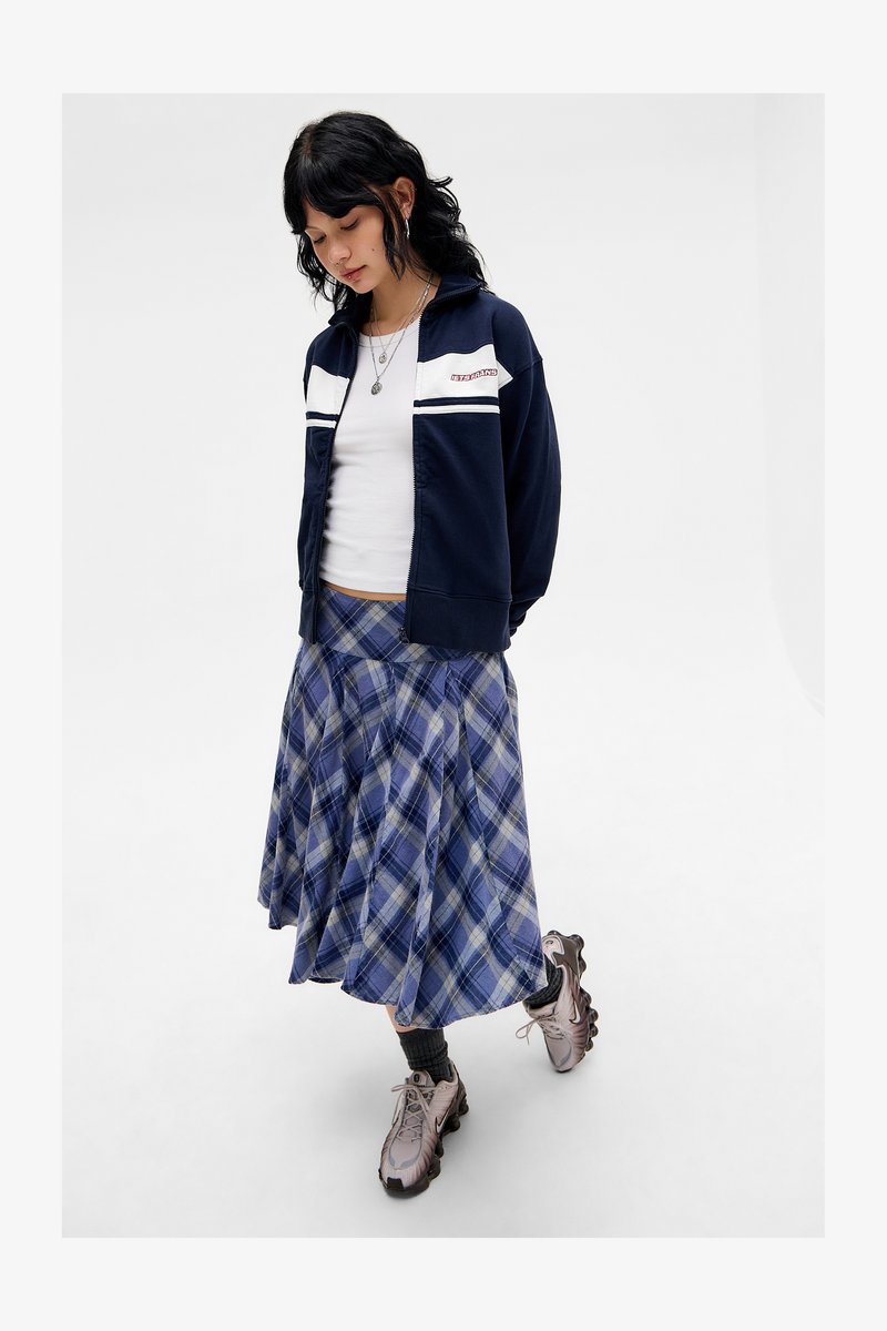 Chaqueta navy con cremallera y rayas blancas, camisa blanca, falda de cuadros azul, calcetines grises y zapatillas gris claro. Tejido suave, patrón juguetón.