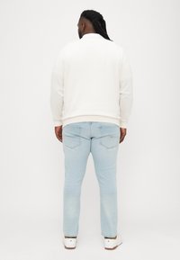 Krém színű pulóver kerek nyakkal, világoskék farmernadrággal, hátul zsebekkel, fehér cipővel viselve. Hátsó nézet.