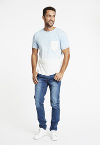 Camiseta de color azul claro con degradado y mangas cortas, con un bolsillo blanco en el pecho, combinada con jeans azules desgastados y zapatillas blancas.