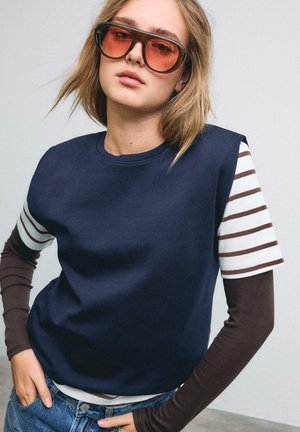 Young woman wearing oversized orange-tinted sunglasses, navy sleeveless top over striped brown and white long sleeves, with hands in jeans pockets.