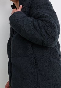 Tmavá fleecová bunda s texturovaným materiálem, vysokým límcem a zapínáním na zip. Má boční kapsy a volný, teplý střih.