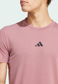 T-shirt rose clair à manches courtes en tissu doux avec un col rond. Présente un logo à trois rayures verticales noires sur la poitrine.