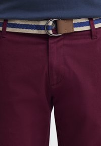 Pantalons bordeaux avec une texture lisse, dotés d'une ceinture tissée beige avec des rayures bleues et une boucle en métal. Coutures détaillées le long des coutures.