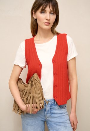 Donna che indossa un gilet rosso con bottoni sopra una t-shirt bianca e jeans blu, che tiene una borsa in pelle scamosciata color cuoio con frange su uno sfondo neutro.