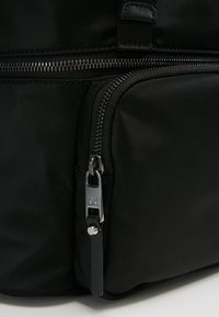 Sac en tissu noir avec deux compartiments zippés visibles, tirette argentée avec logo "MK" et languette en cuir sur la petite poche.