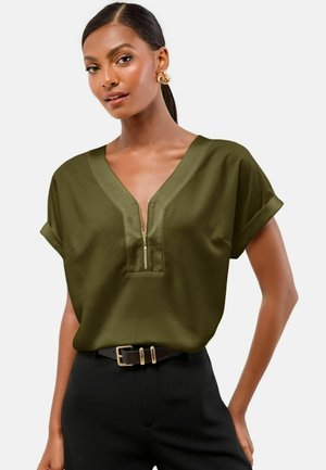 Woman wearing an olive green short-sleeve blouse with a front zipper and black pants with a black belt, standing against a plain background.